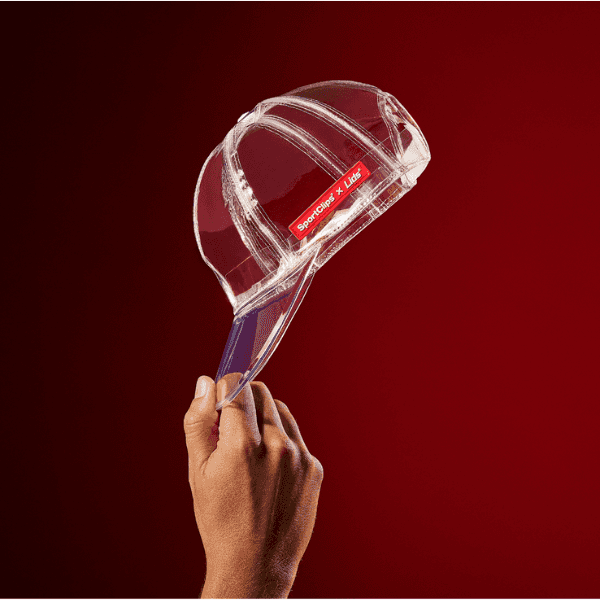 Sport Clips x Lids Clear Hat collaboration showcasing a unique transparent cap design, held by a hand against a bold red background.