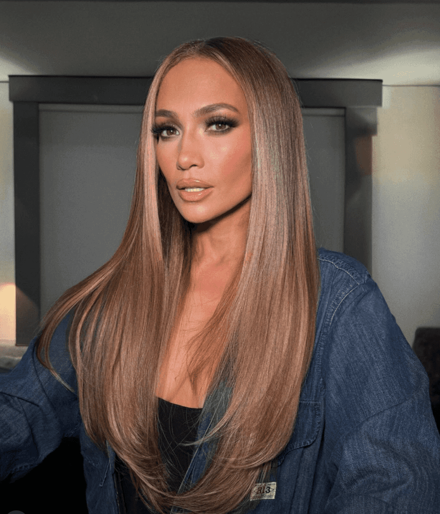 Jennifer Lopez with long, sleek honey-blonde hair styled straight with a center part, wearing a dark denim jacket and neutral glam makeup, sitting in a softly lit indoor setting.