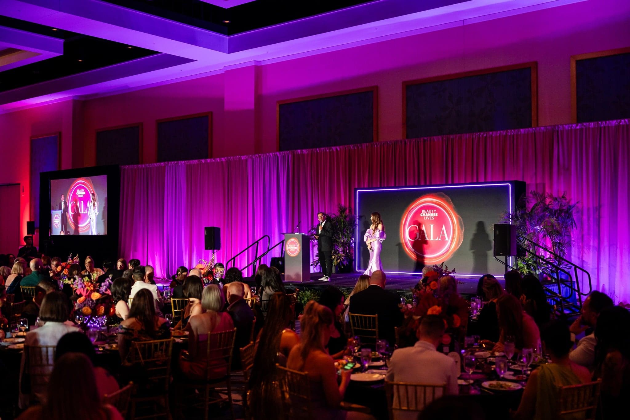 beauty changes lives 2025 gala with tables and stage with hosts sam villa and elizabeth faye addressing the crowd