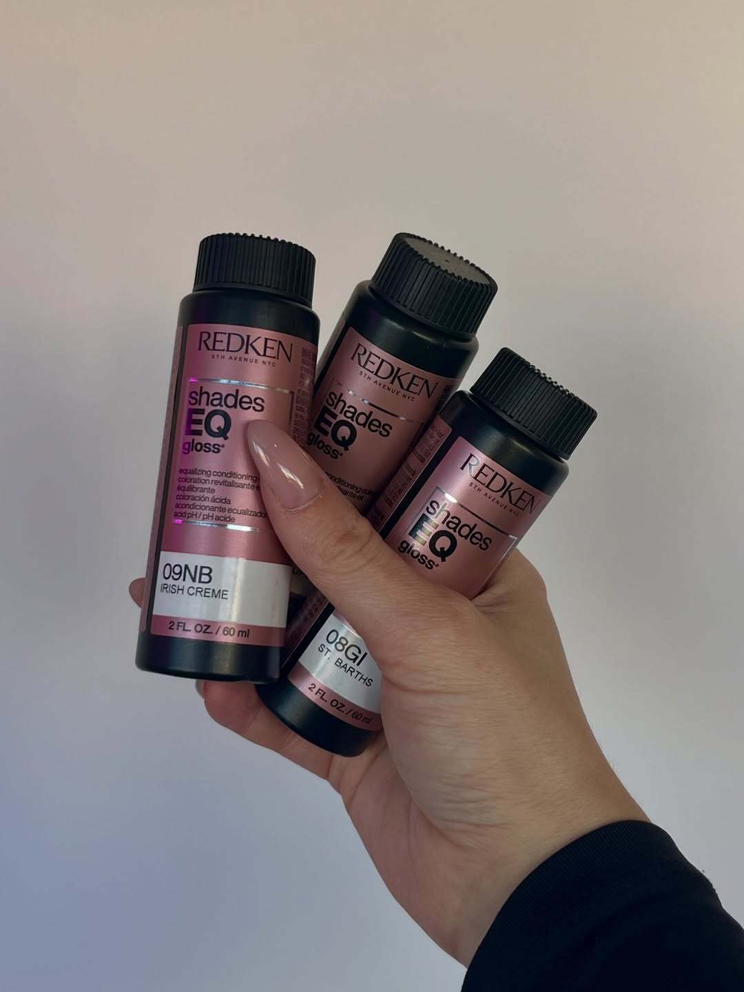 Hand holding three bottles of Redken Shades EQ Gloss hair color, including shades 09NB Irish Crème and 08GI St. Barths, against a plain light background. The bottles feature the brand’s signature rose-gold labels and black caps.