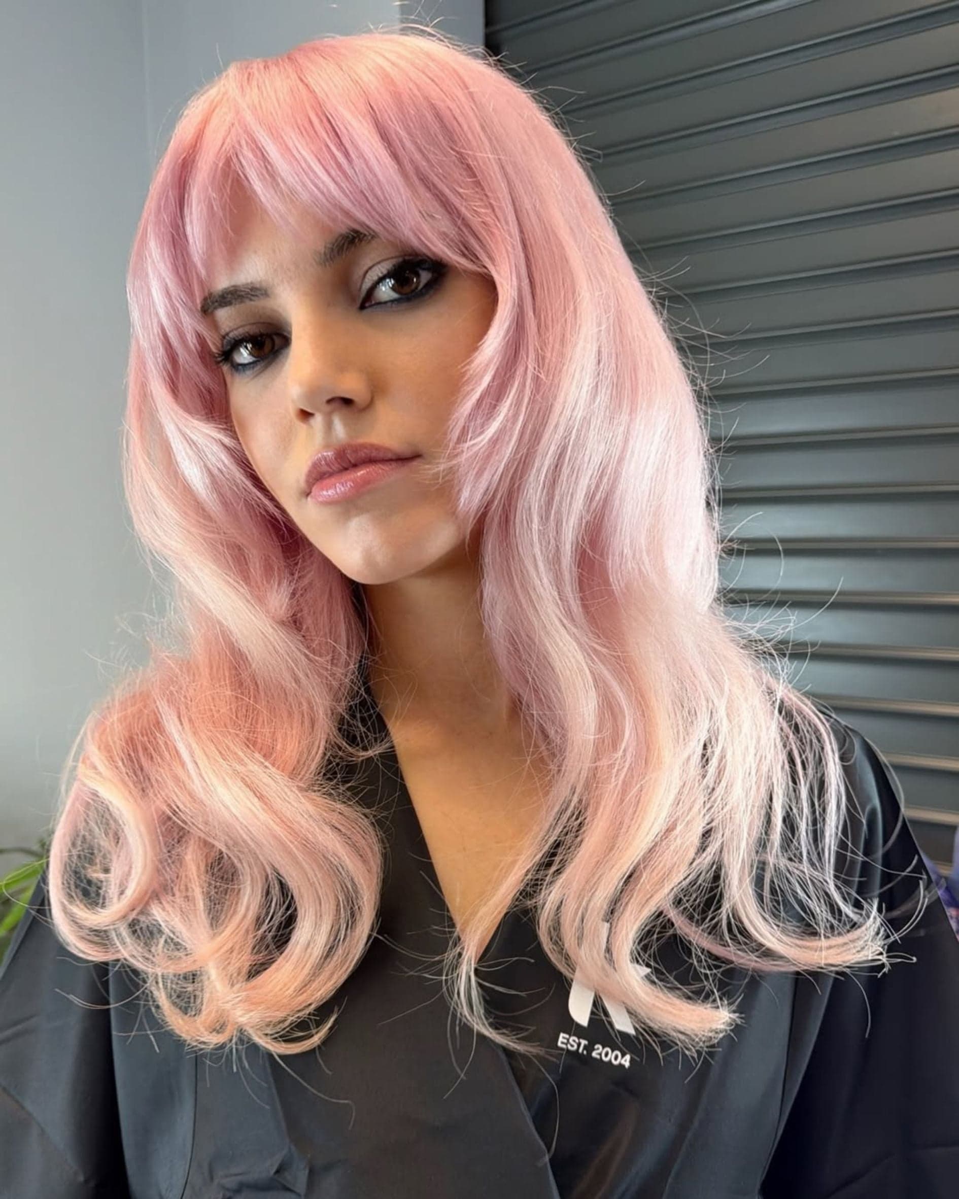 Soft pastel pink hair with long layers and curtain bangs, featuring a sheer, wearable pink tone created on a light blonde base.