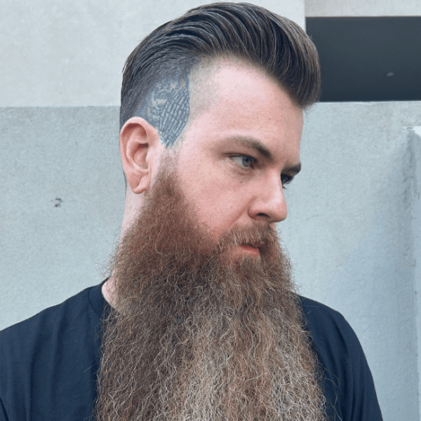 A man in a black shirt showcases a men's haircut and beard styling by Bertus, featuring a clean fade, slicked-back hair, and a full beard.