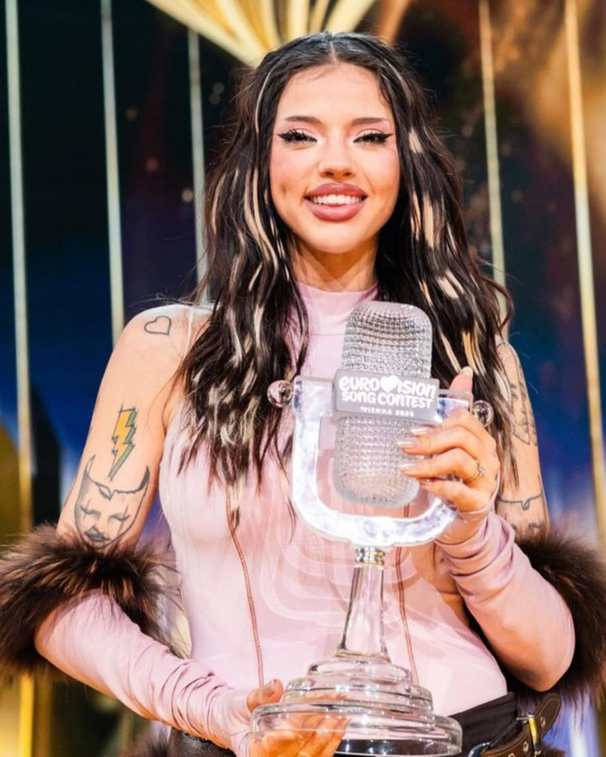 A Eurovision artist poses with the Eurovision trophy backstage featuring dimensional brunette hair with bold face-framing highlights.
