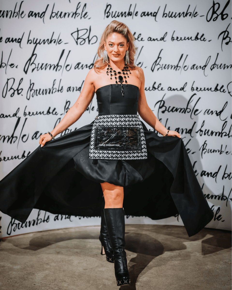 Ashlee Norman poses in front of a Bumble and bumble backdrop wearing a black dress, statement jewelry and boots while holding out the sides of her skirt.