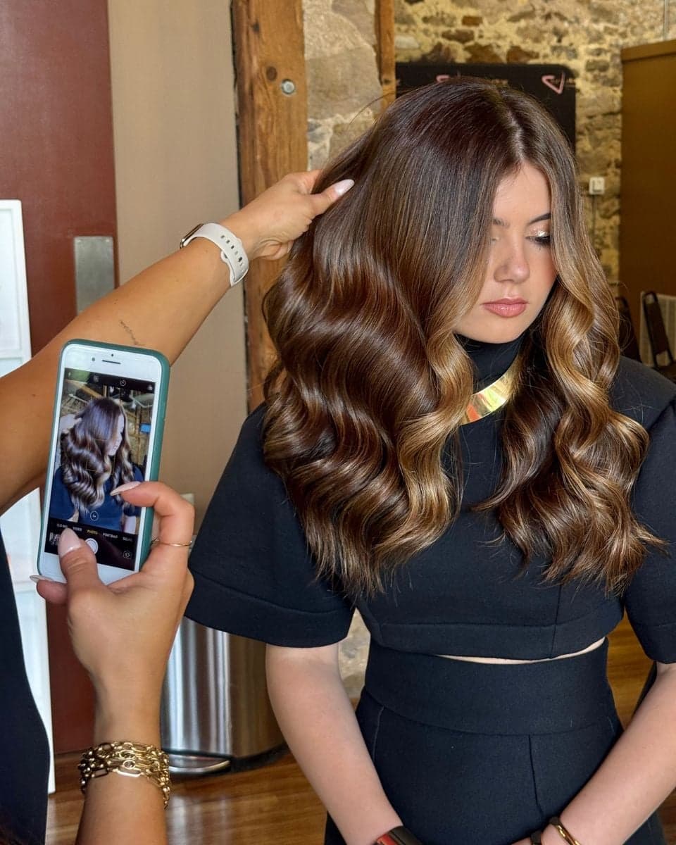 A brunette client with long, glossy waves showcases a luxury balayage color featuring rich chocolate brown tones blended with soft caramel ribbons. A stylist lifts the hair to display dimension while another photographs the finished result on a smartphone inside a salon.