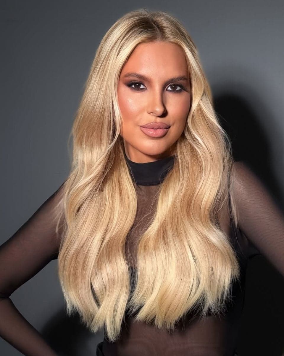 Portrait of long champagne blonde hair with soft beige dimension, a seamless root blend and polished waves styled over a sheer black top.