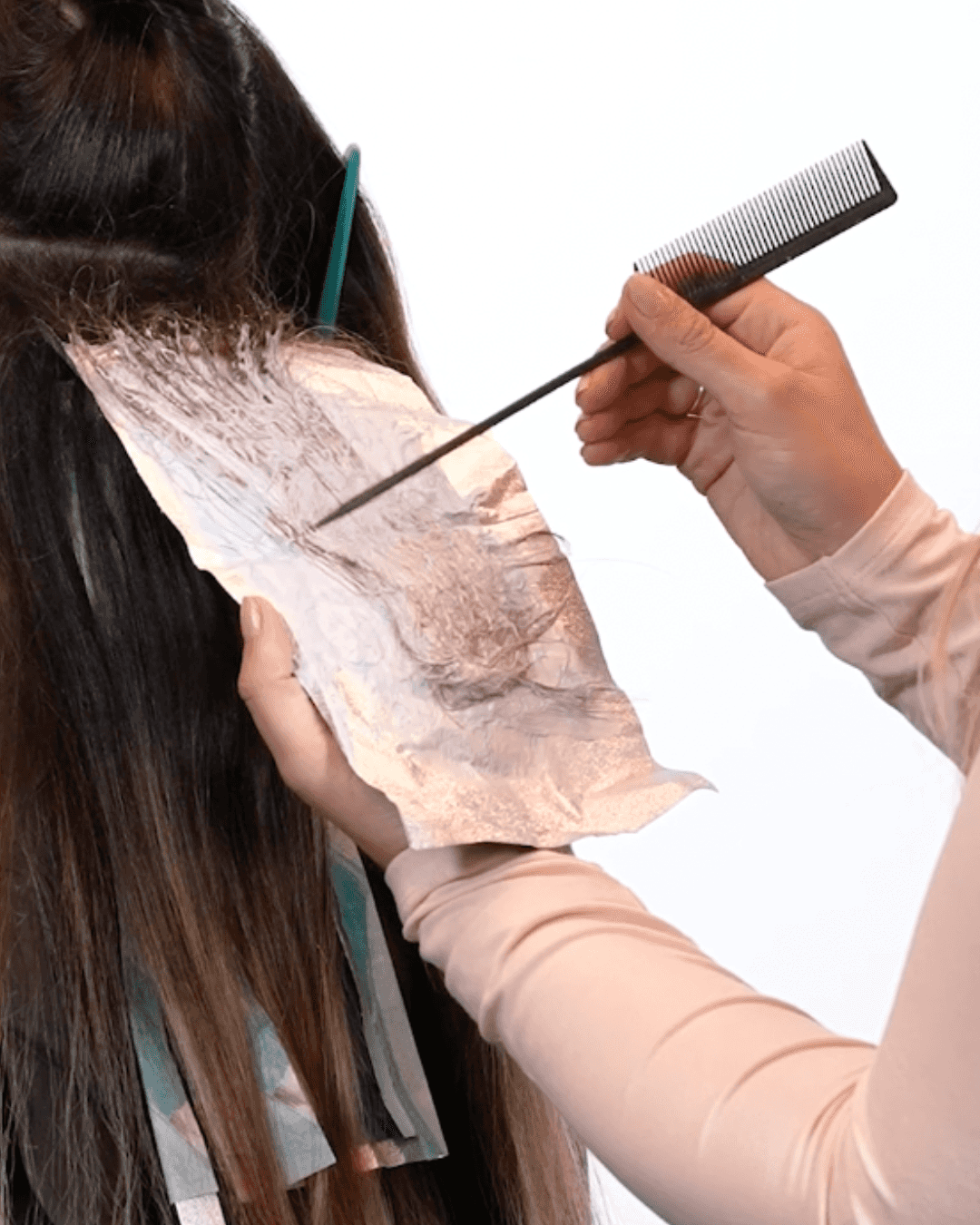 A stylist holds an open foil while using the tail of a comb to smooth and separate hair strands coated with lightener. The technique demonstrates how to correct hair that has folded or shifted inside the foil before closing it.