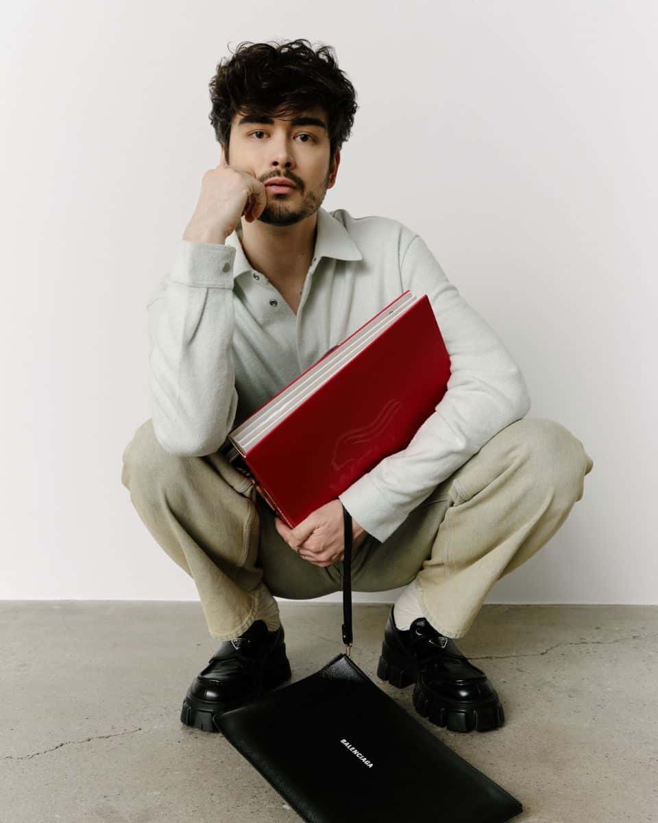 Hairstylist Chris Weber poses holding a red Wella portfolio with a black Balenciaga bag in front of him.